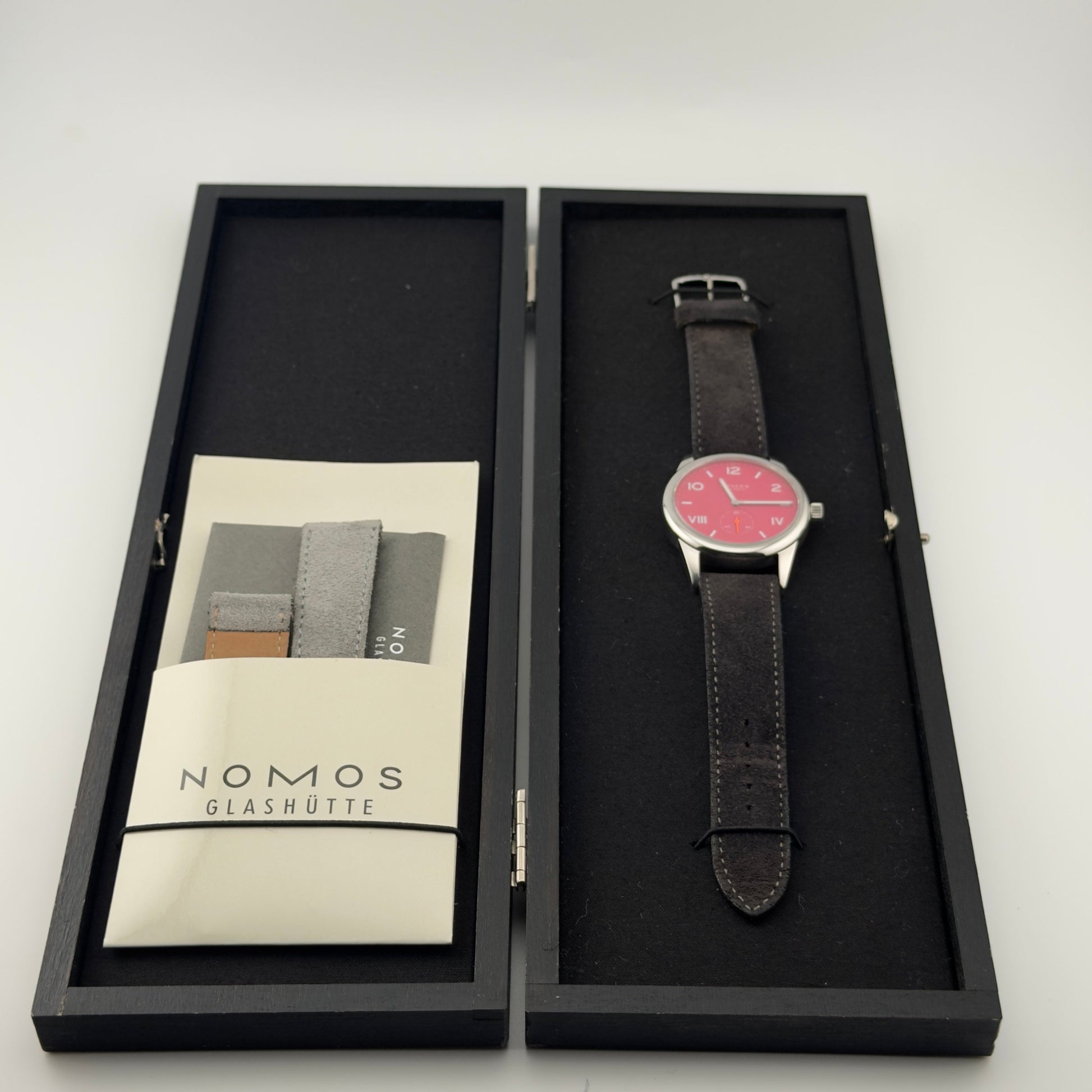 Watch with black leather strap and red face in a Nomos Glashütte box on a white background